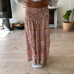 Färgglad långkjol med paisleymönster - Superfin långkjol med färgstarkt paisleymönster i rosa, orange, beige och inslag av blått. Kjolen har smock i midjan för skön passform och är gjord i ett lätt, flowy material som känns perfekt för varmare dagar. Snygg att styla med en enkel topp och sandaler.