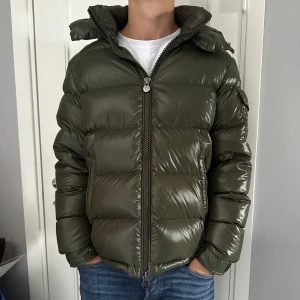 Olivgrön Moncler pufferjacka - Säljer en olivgrön pufferjacka från Moncler med glansig finish och klassisk logga på ärmen. Perfekt för dig som vill ha en jacka för vintern! Jackan är äkta och i nyskick därav priset!