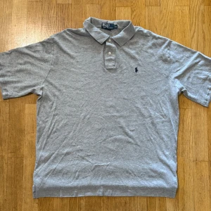Grå pikétröja från Ralph Lauren XL - Klassisk grå pikétröja från Ralph Lauren i storlek XL. Tröjan har korta ärmar, krage med två knappar och den ikoniska broderade loggan på bröstet. Tillverkad i mjuk bomull som känns skön mot huden. Perfekt för en clean och stilren look.