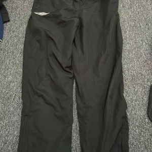 Svarta täckbyxor med dragkedja - Svarta täckbyxor med elastisk midja och dragkedja längs benen. Perfekta för kalla dagar och utomhusaktiviteter. Materialet är slitstarkt och vattenavvisande, med en enkel design och diskret logga på ena benet. Pris kan diskuteras 😊