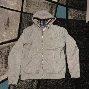 Grå hoodie från Burberry med dragkedja - Grå hoodie från Burberry i bomull med klassiskt rutigt foder i huvan. Tröjan har hel dragkedja, broderad logga på bröstet och två fickor framtill. Perfekt för dig som gillar stilren och lyxig streetwear.