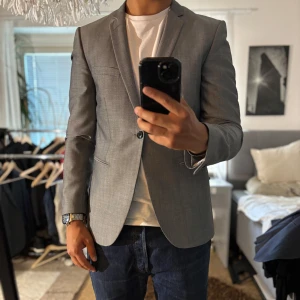 Grå kavaj från Bläck slim fit - Snygg grå kavaj från Bläck i slim fit-modell. Klassisk design med smala slag, två framfickor och en bröstficka. Perfekt för dig som gillar stilrena och moderna plagg. Fodrad insida med svart tyg och diskreta detaljer.
