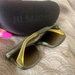 Jil Sander solglasögon khaki/gul - Snygga solglasögon från Jil Sander med transparent grå båge och coola gula detaljer på insidan av skalmarna. Rektangulär form och tonade linser ger en modern vibe. Modell JS735S, tillverkade i Italien. Perfekta för dig som vill sticka ut. Äkta och kommer med etui dock utan trasa. Vet inte vart den är 