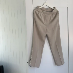 Beiga kostymbyxor från NA-KD, stl 36 - Snygga beiga kostymbyxor från NA-KD i rak modell med pressveck och hög midja. Byxorna har diskreta fickor bak och är tillverkade i ett mjukt, vävt material som ger en clean och stilren look. Perfekta att matcha med en enkel topp eller kavaj.