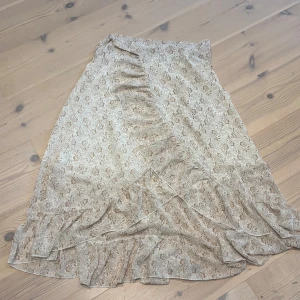 Beige blommig omlottkjol med volang - Supersöt långkjol i beige med blommigt mönster och omlottmodell. Kjolen har fina volangdetaljer längs kanten och är perfekt för en romantisk look. Lätt och flowigt material som känns luftigt.