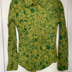 Grön blommig skjorta från Indiska - Supersnygg grön skjorta från Indiska med blommigt mönster i mörkare grönt. Skjortan har klassisk krage, två bröstfickor med lock och knappar framtill samt vid ärmslut. Figurnära passform och mjukt bomullsmaterial. Perfekt för dig som gillar retro vibes!