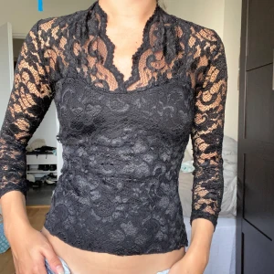 Svart spetsblus med trekvarts ärm - Supersnygg svart blus i spets med lång ärm och v-ringad hals. Transparent upptill och på ärmarna, med täckt bröstparti. Perfekt för dig som vill ha en elegant och trendig look med coola detaljer.