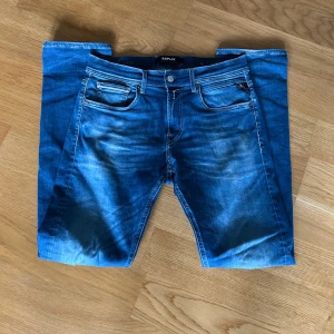 Blå Replay Anbass jeans 30/32 - Snygga blå Replay Anbass jeans med slim passform och klassisk femficksdesign med storlek 30/32. Jeansen har slitningar och en mindre reva på ena benet. Tydliga ljusa partier framtill och bakficka med broderad detalj. Tveka inte över att ställa frågor