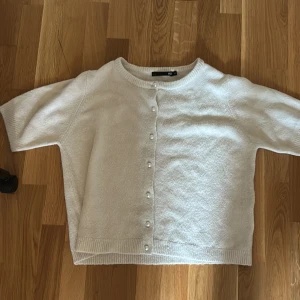Vit kofta med pärlknappar från 157 - Superfin vit kofta från 157 med kort ärm och pärlknappar framtill. Koftan har rund halsringning och ribbade muddar vid ärmslut och nederkant. Perfekt att slänga över en topp för en clean och söt look.