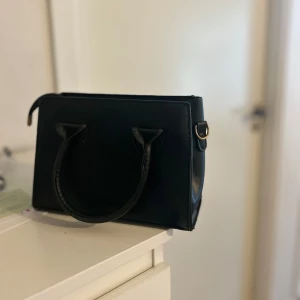 Svart stilren handväska i skinnimitation - Säljer en svart, fyrkantig handväska med två rejäla handtag och guldfärgade metalldetaljer. Väskan har en minimalistisk design och är tillverkad i skinnimitation. Perfekt för dig som gillar en clean och klassisk look.