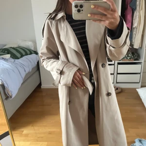 Beige trenchcoat från ADPT - Säljer en beige trenchcoat från ADPT med klassisk dubbelknäppning, bälte i midjan och snygga detaljer vid ärmsluten. Jackan är aldrig använd!