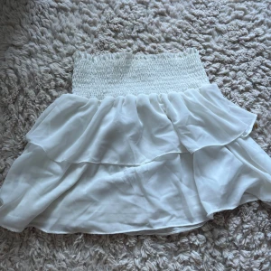 Vit volangkjol från Chelsea - Supersöt vit kortkjol från Chelsea med bred smock i midjan och dubbla volanger. Kjolen är luftig och har en flowy känsla, perfekt för varma dagar. Tillverkad i ett lätt material som ger en romantisk vibe.