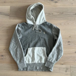 Grå hoodie från Polo Ralph Lauren - Snygg tvåfärgad hoodie från Polo Ralph Lauren i grått och ljusgrått. Klassisk huva med snörning, känguruficka och broderad logga på bröstet. Mjuk bomullskvalitet och ribbade muddar. Perfekt för chill dagar eller när du vill vara casual. Är lite urtvättad men inget större problem