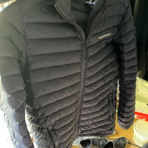 Marin blå dunjacka Peak Performance - Säljer en marin blå dunjacka från Peak Performance med horisontella sömmar och dragkedja framtill. Jackan har en clean look, hög krage och huva. Perfekt för kyliga dagar och enkel att matcha med allt. Klassisk och stilren design.