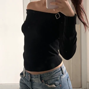 Svart offshoulder stickad topp - Snygg svart stickad topp med offshoulder-design och lång ärm. Toppen har en cool detalj med spänne på ena axeln och ribbad kant upptill. Perfekt att styla med jeans för en trendig look.