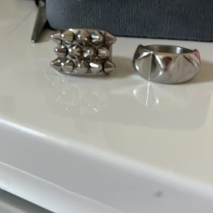 Två silvriga Edblad ringar med nitar - Säljer två coola ringar från Edblad i blankt silverfärgat metall. Ena ringen har flera små nitar runt hela bandet, medan den andra har större, spetsiga detaljer. Strlk 17.5 på båda. De kostar 300 tillsammans.