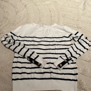 Vit och marinblå randig tröja CC Fashion - Snygg vit stickad tröja med marinblå ränder från CC Fashion. Långärmad modell med ribbade muddar och dekorativa guldknappar vid ärmsluten. Perfekt för dig som gillar klassisk stil med en twist. Passar till både jeans och kjol.