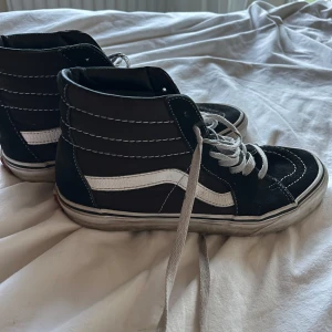 Svarta Vans Sk8-Hi sneakers - Klassiska svarta Vans Sk8-Hi sneakers med vit sula och vit sidostrip. Högt skaft, vit snörning och kontrastsömmar. Ovandel i mocka och canvas för en cool streetstil. Perfekta för dig som gillar skate eller vill ha en tidlös look.