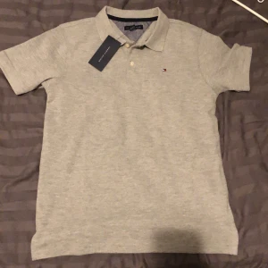 Grå pikétröja från Tommy Hilfiger - Helt ny grå piké köptes i USA för ungefär 800kr och säljer den för 600kr. Pris kan diskuteras vid snabb affär. Passar någon på 160cm. (M för barn)