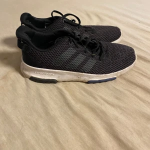 Svarta Adidas sneakers med Cloudfoam - Svarta Adidas sneakers med klassiska tre ränder på sidan och vit Cloudfoam-sula. Ovandelen är i mesh för extra andningsförmåga och skon har snörning. Perfekta för dig som gillar sportig och avslappnad stil.