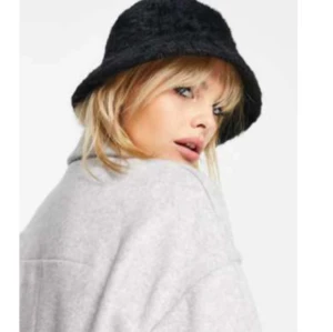 Svart bucket hat i teddy - Trendig svart bucket hat i mjuk teddy-look. Perfekt accessoar för att lyfta din outfit och hålla värmen. Klassisk form med nedvikt brätte och fluffig känsla som ger en cool vibe.