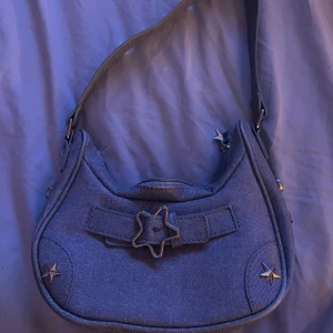 Y2k insperad axelväska  - Söt och stilren liten axelväska i ljusblå färg. Gjord i plastmaterial men med en jeansliknande look som ger den en trendig touch. Väskan är dekorerad med charmiga stjärndetaljer i metall och en stjärnformad spänne framtill. Perfekt för utekvällen, konserten eller en dag på stan – liten och smidig men rymmer det viktigaste som mobil, nycklar och plånbok.  🌟 Justerbar rem 🌟 Dragkedja upptill 🌟 Tygfoder invändigt 🌟 Unik design med stjärntema  Använd endast ett fåtal gånger – i mycket fint skick!
