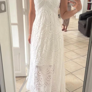 Vit långklänning med broderade detaljer - Superfin vit långklänning med broderade hålmönster och v-ringad hals. Klänningen är ärmlös och har en luftig känsla, perfekt för varma dagar. Snygg passform och romantiska detaljer som ger en bohemisk vibe.