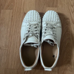 Louboutin  - Unika vita sneakers från Christian Louboutin med coola nitar på tån och klassisk röd sula. Skorna har snörning och är tillverkade i skinn. Perfekta för dig som vill sticka ut med en edgy och lyxig stil.