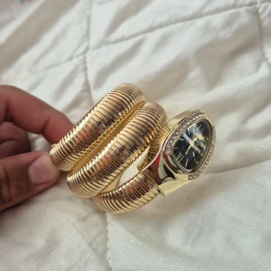 Guldig ormformad klocka - Säljer en unik guldig klocka med spiralarmband som liknar en orm. Urtavlan är svart med romerska siffror och omges av gnistrande stenar. Klockan har en cool, modern look och passar dig som vill sticka ut med accessoarer.