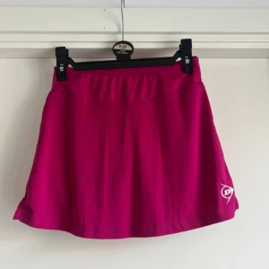 Rosa sportkjol från Dunlop - Snygg rosa sportkjol / skort / kjol med shorts från Dunlop med elastisk midja och diskret logga framtill. Kjolen är kort och tillverkad i ett lätt och stretchigt syntetmaterial som passar perfekt för tennis eller andra sportiga aktiviteter.