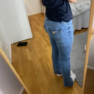 Ljusblå bootcut jeans med fickdetalj - Säljer ett par ljusblå jeans med bootcut-modell och snygga guldknappar på bakfickorna. Jeansen har klassisk femficksdesign och är tillverkade i mjukt denimtyg med lite stretch. Perfekta för dig som gillar en retro vibe och vill ha en bekväm passform.