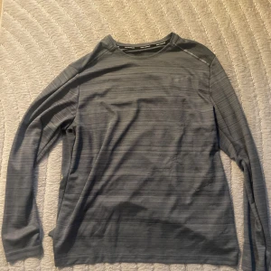 Grå långärmad tröja från Nike - Säljer en grå långärmad tröja från Nike med diskret randigt mönster. Tröjan har en klassisk passform och är tillverkad i ett mjukt och lätt material som passar perfekt för lager-på-lager. Enkel och stilren design för en clean look.