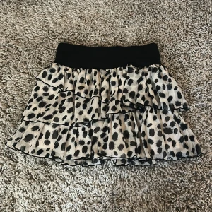 Prickig volangkjol med resårmidja - Supersöt kortkjol med svart resårmidja och flera lager volanger. Kjolen är vit med stora svarta prickar och har en luftig, flowy känsla. Perfekt för dig som gillar att sticka ut med ett lekfullt mönster och voluminös look.