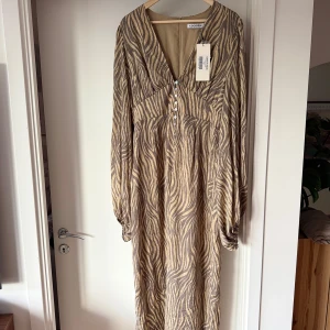 Zebramönstrad långklänning från Adoore - Snygg långklänning från Adoore med zebramönster i beige och brunt. Klänningen har v-ringning, knappar framtill och långa ärmar med lätt puff. Perfekt för dig som vill sticka ut med ett trendigt djurmönster. Ligger ute på fler sidor där av har jag valt att ta bort köp via Plick:)