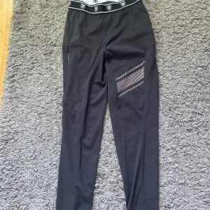 Svarta Fairfield ridleggings S - Svarta ridleggings från Fairfield med bred elastisk midja med logga, silikonmönster för extra grepp och praktisk ficka på sidan. Snygga detaljer med vita ränder och diskret mönster på benen. Perfekta för ridning och träning. Pris kan diskuteras.