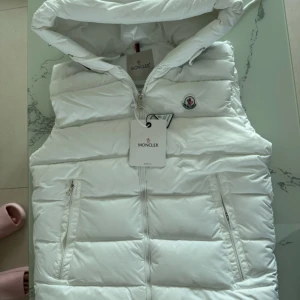Vit dunväst från Moncler med huva - Snygg vit dunväst från Moncler med huva och dragkedja framtill. Västen har två fickor med dragkedja och klassisk Moncler-logga på bröstet. Quiltad design och sportig look, perfekt för lager-på-lager-stil. Insidan har en cool serietidningsdetalj. nyköpt men säljer för den ej passar mig den passar folk runt 160-172cm 