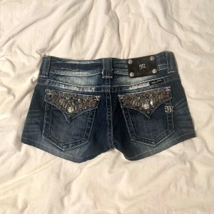 Miss Me jeansshorts med bling, stl 26 - Snygga mörkblå jeansshorts från Miss Me med blingade bakfickor. Shortsen har låg midja, dekorativa nitar och stenar på fickorna samt tydliga kontrastsömmar. Perfekta för dig som gillar detaljer och vill sticka ut i sommar. Midjemått: ca 38.5 cm