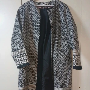 Svartvit mönstrad rock/kappa - Snygg svartvit kappa/rock med grafiskt mönster och kontrasterande detaljer vid ärmslut och nederkant. Rak passform, rund halsringning och helsvart foder. 