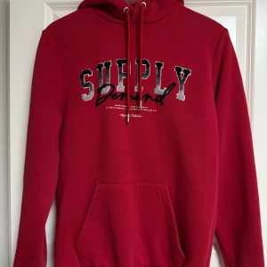 Röd hoodie från Supply & Demand XS - Röd hoodie från Supply & Demand med broderad text på bröstet. Klassisk känguruficka framtill, justerbar huva med snören och mjukt fleecetyg på insidan. Perfekt för dig som gillar streetwear och vill ha en snygg och bekväm look.