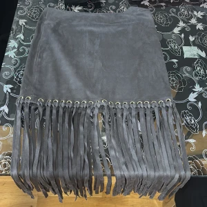 Grå mockakjol med fransar Gina Tricot - Supersnygg grå kortkjol i mockaimitation från Gina Tricot. Kjolen har coola fransdetaljer längs nederkanten och metallringar som ger en edgy vibe. Perfekt för dig som vill sticka ut med en trendig look.