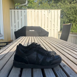 Svarta Valentino Rockrunner sneakers - Snygga helsvarta Valentino Rockrunner sneakers med coola nitar på hälen. Skorna har en mix av skinn och mocka, klassisk snörning och platt sula. Perfekta för dig som gillar en stilren och edgy look. 