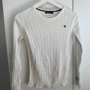 Vit kabelstickad tröja Gina Tricot - Vit kabelstickad tröja från Gina Tricot med rund halsringning och diskret blå brodyr på bröstet. Tröjan har långa ärmar och ribbade muddar vid ärmslut och nederkant. Perfekt för en clean och klassisk look.