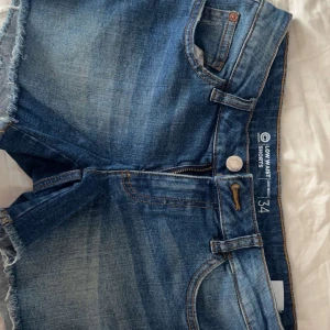 Blå jeansshorts low waist - Snygga blå jeansshorts med låg midja och råa kanter. Klassisk femficksmodell med dragkedja och knapp framtill. Perfekta för sommardagar och enkel att matcha med allt.