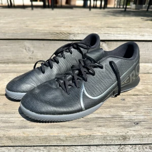 Svarta Nike fotbollsskor med grå sula - Snygga svarta Nike fotbollsskor med grå sula och diskret Nike-logga på sidan och undersidan. Skorna har snörning och är tillverkade i syntetmaterial med texturerad yta för bättre bollkontroll. Perfekta för inomhusspel och har en låg profil.