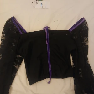 Svart topp med spetsärmar och lila band - Unik svart topp med transparenta spetsärmar och lila sammetsband längs axlarna och framtill. Offshoulder-modell som ger en cool och edgy vibe. Perfekt för dig som vill sticka ut med detaljer och kontraster.