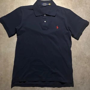 Marinblå pikétröja från Ralph Lauren - Snygg marinblå pikétröja från Polo Ralph Lauren i slim fit-modell. Klassisk krage, korta ärmar och knäppning med två knappar. Ikoniska röda loggan broderad på bröstet. Tillverkad i mjuk bomull som känns skön mot huden. Hör av vid minsta fundering!💫