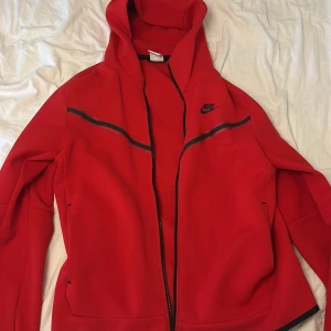 Röd Nike hoodiejacka med dragkedja - Nike hoodiejacka i starkt röd färg med svarta detaljer och dragkedja hela vägen. Jackan har huva, långa ärmar och sportig look. Perfekt för chill dagar eller när du vill ha en clean streetstyle. Snygg Nike-logga på bröstet och praktiska fickor på sidorna.