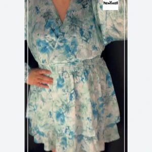 Blommig kortklänning från New Yorker - Supersöt kortklänning med blått och vitt blommönster från New Yorker. Klänningen har omlottliv, markerad midja med resår och volangig kjol. Långa ärmar och lätt, luftig känsla – perfekt för varma dagar.