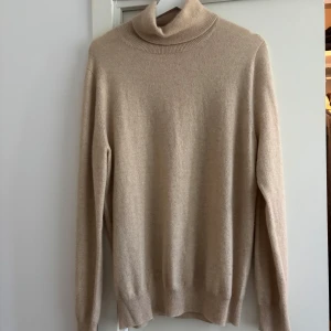 Beige polotröja i kashmir från Gobi - Supermjuk beige polotröja från Gobi i 100% kashmir. Klassisk modell med ribbade muddar och hög polokrage. Perfekt för dig som gillar en clean och stilren look med lyxig känsla. Sitter normalt och är långärmad.