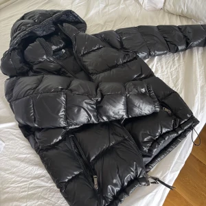 Svart pufferjacka Polo Ralph Lauren - Säljer en svart, glansig pufferjacka från Polo Ralph Lauren. Jackan har huva, tvåvägsdragkedja med Polo-detaljer och stora quiltade partier. Perfekt för kalla dagar och riktigt snygg till streetwear-looken.